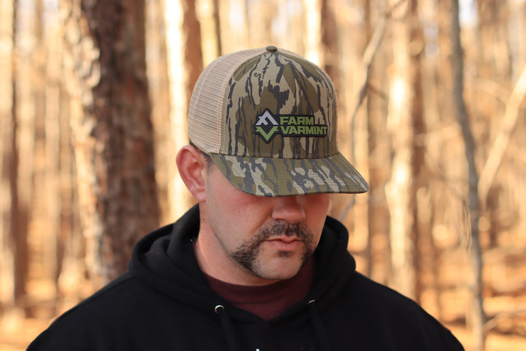 Man wearing a camouflage cap with 'FarmVarmint' logo in a forest setting